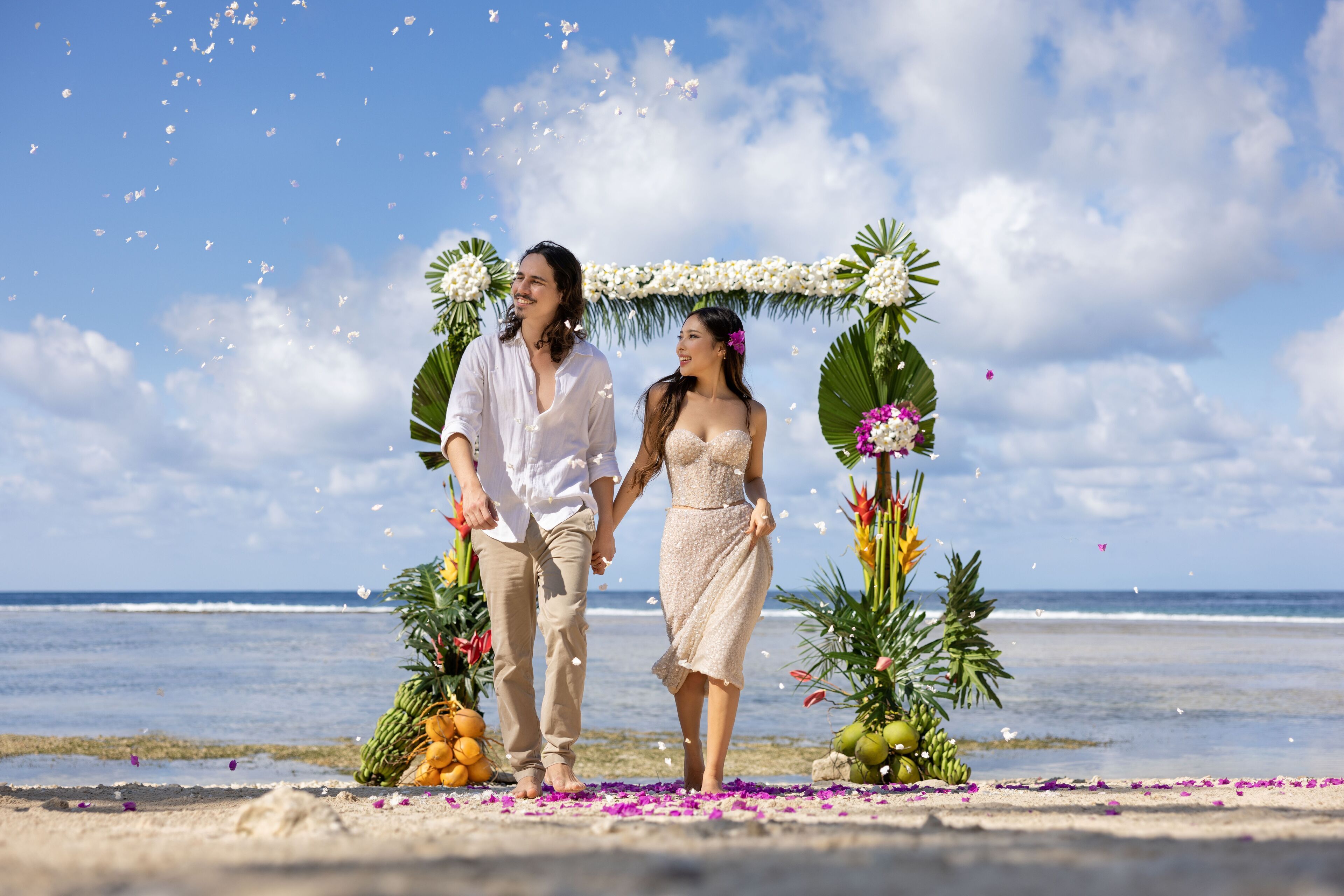 Una pareja tomándose de las manos bajo un arco floral en una playa, con el océano al fondo.
