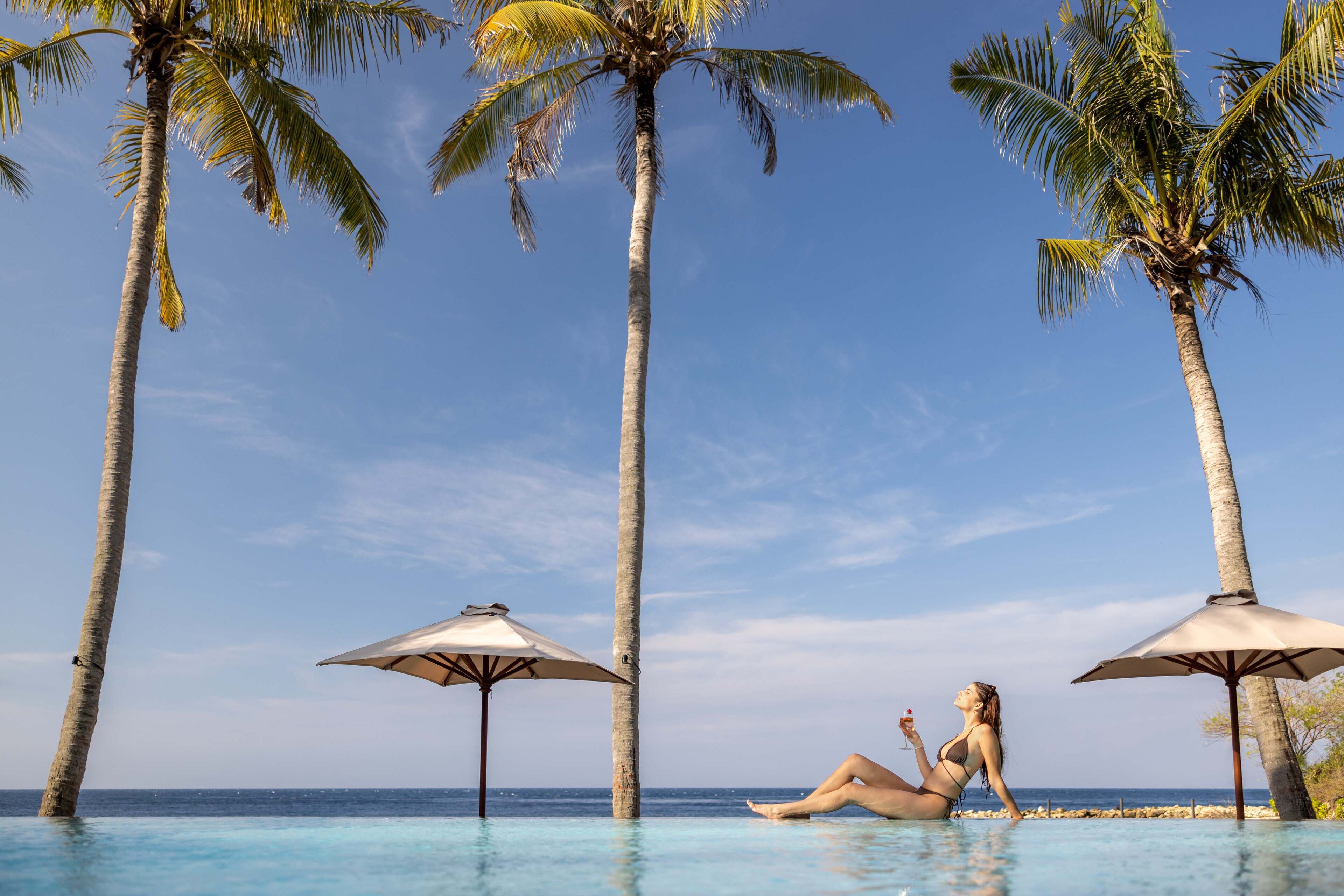 A woman relaxing in a pool with palm trees and umbrellas.