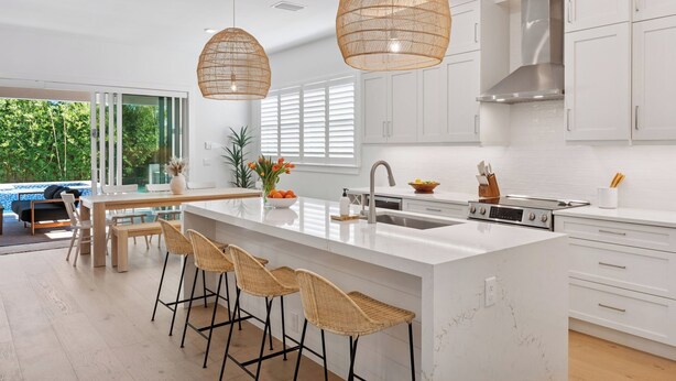 Une cuisine moderne avec des meubles blancs, un grand îlot central doté de tabourets, des luminaires suspendus et une vue sur une piscine à l’extérieur.