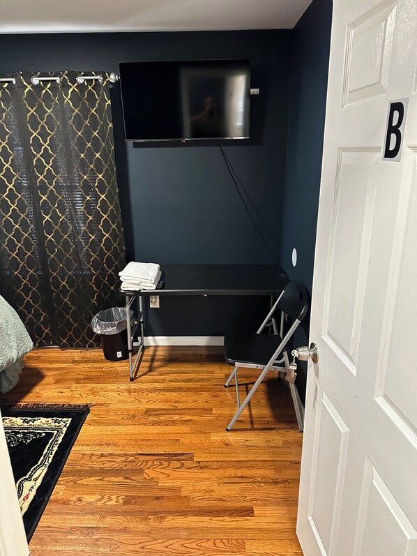 A compact room with a bed, a desk, a chair, a TV mounted on the wall, and a door marked with a letter.