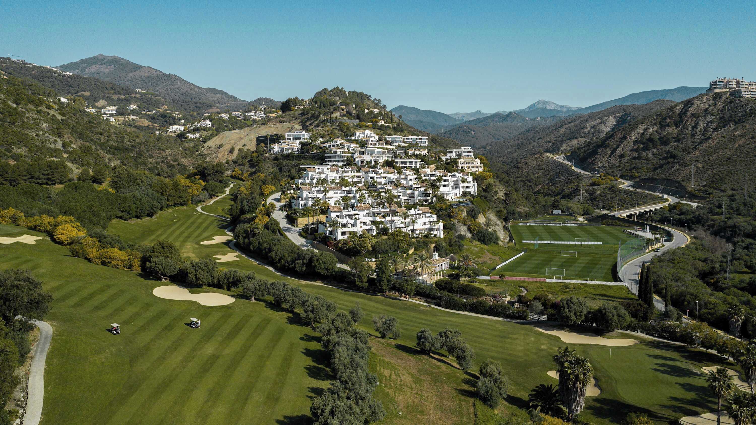 An aerial view of a golf course with a cluster of white buildings on a hillside, surrounded by trees and mountains.