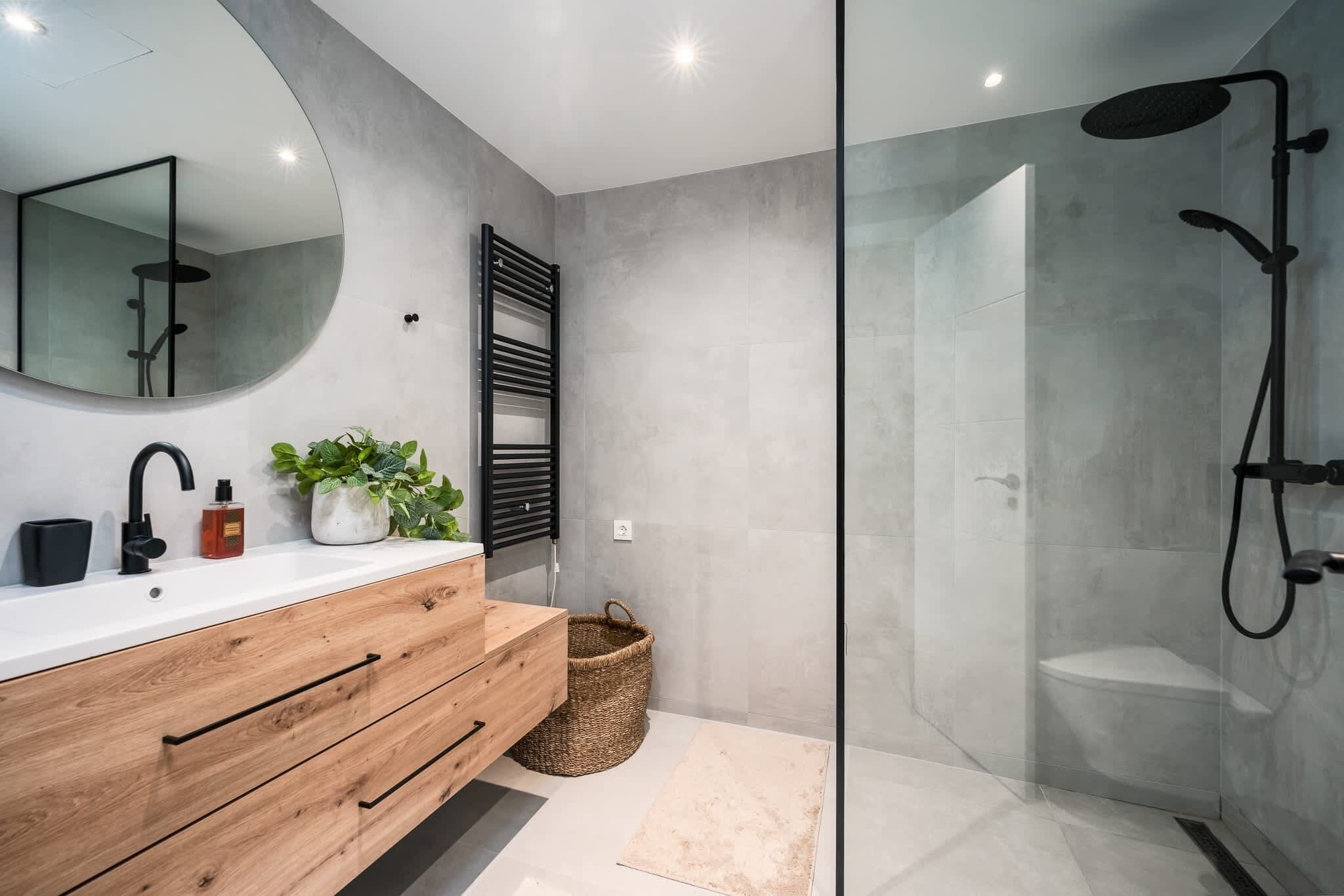 Une salle de bain moderne avec un meuble-lavabo en bois, un miroir rond, une douche dotée d’une paroi en verre et une plante en pot.