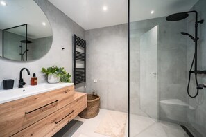 A modern bathroom with a wooden vanity, a round mirror, a shower with a glass enclosure, and a potted plant.