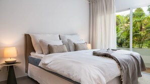 A bedroom with a bed, pillows, a bedside table with a lamp, and a view of greenery outside. - Lomas de La Quinta | Modern Sea View Apt (Benahavís)