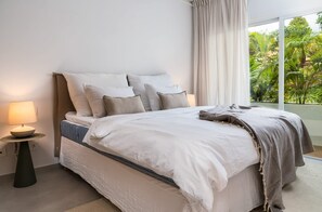 A bedroom with a bed, pillows, a bedside table with a lamp, and a view of greenery outside.