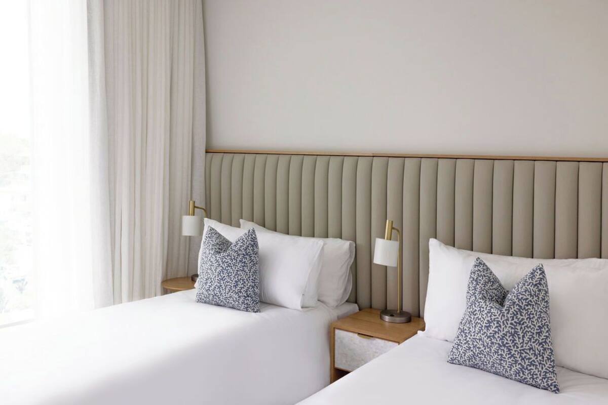 A hotel room with two beds, a nightstand, a lamp, and a window with curtains.