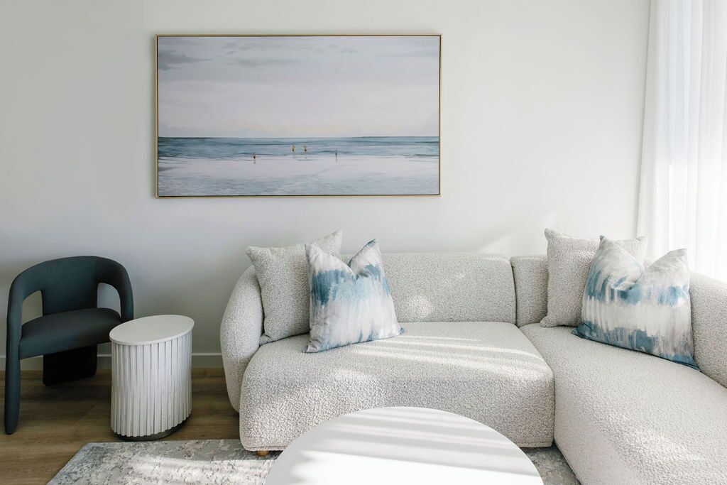 Una sala moderna con un sofá gris, cojines a juego, una mesita redonda blanca, una silla y una obra enmarcada con una escena de playa.
