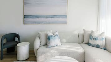 Una sala moderna con un sofá gris, cojines a juego, una mesita redonda blanca, una silla y una obra enmarcada con una escena de playa.