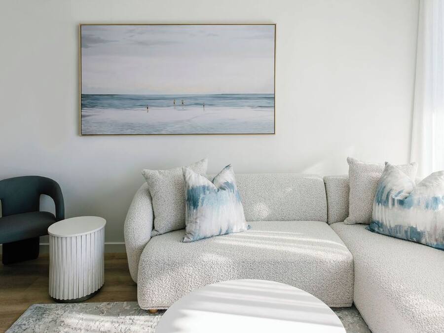 Una sala moderna con un sofá gris, cojines a juego, una mesita redonda blanca, una silla y una obra enmarcada con una escena de playa.