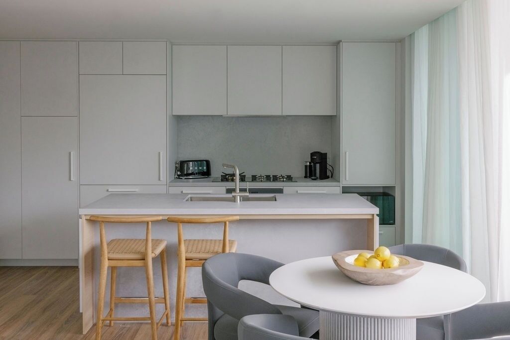 A modern kitchen with a central island, bar stools, and a dining table with a bowl of fruit.