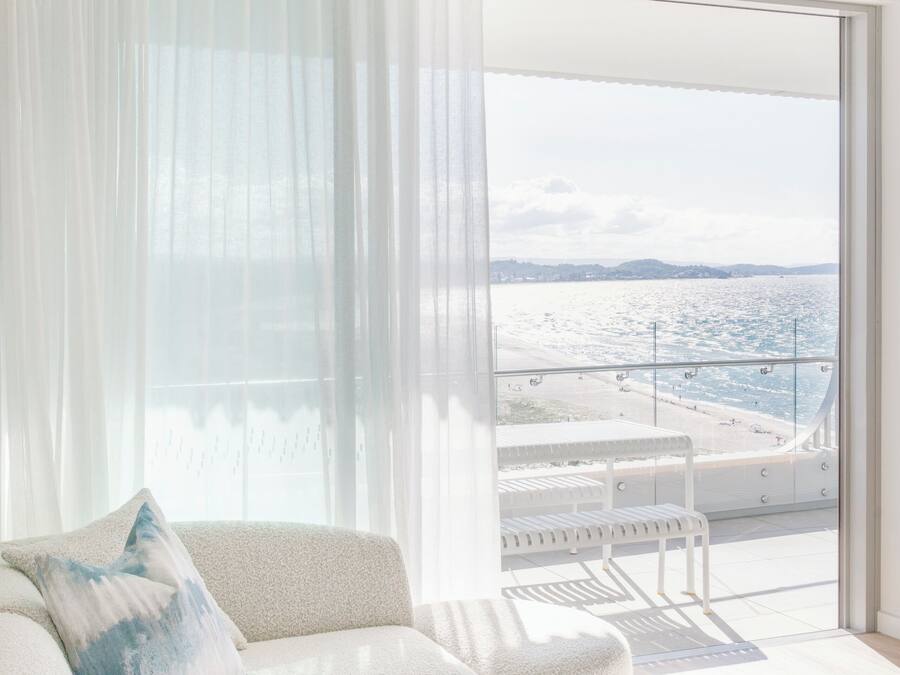 Una sala de estar moderna con un sofá blanco, una mesa redonda y vistas a la playa a través de grandes ventanales.
