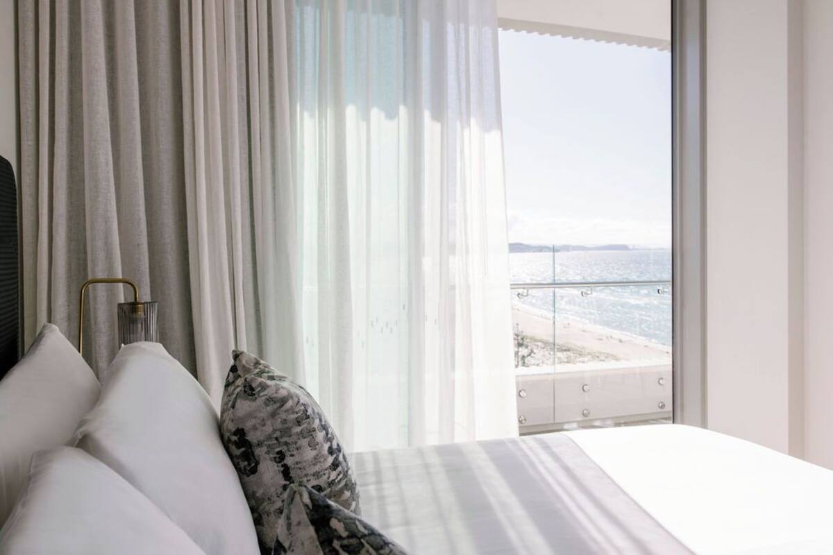 A bedroom with a bed, white curtains, and a view of the beach.