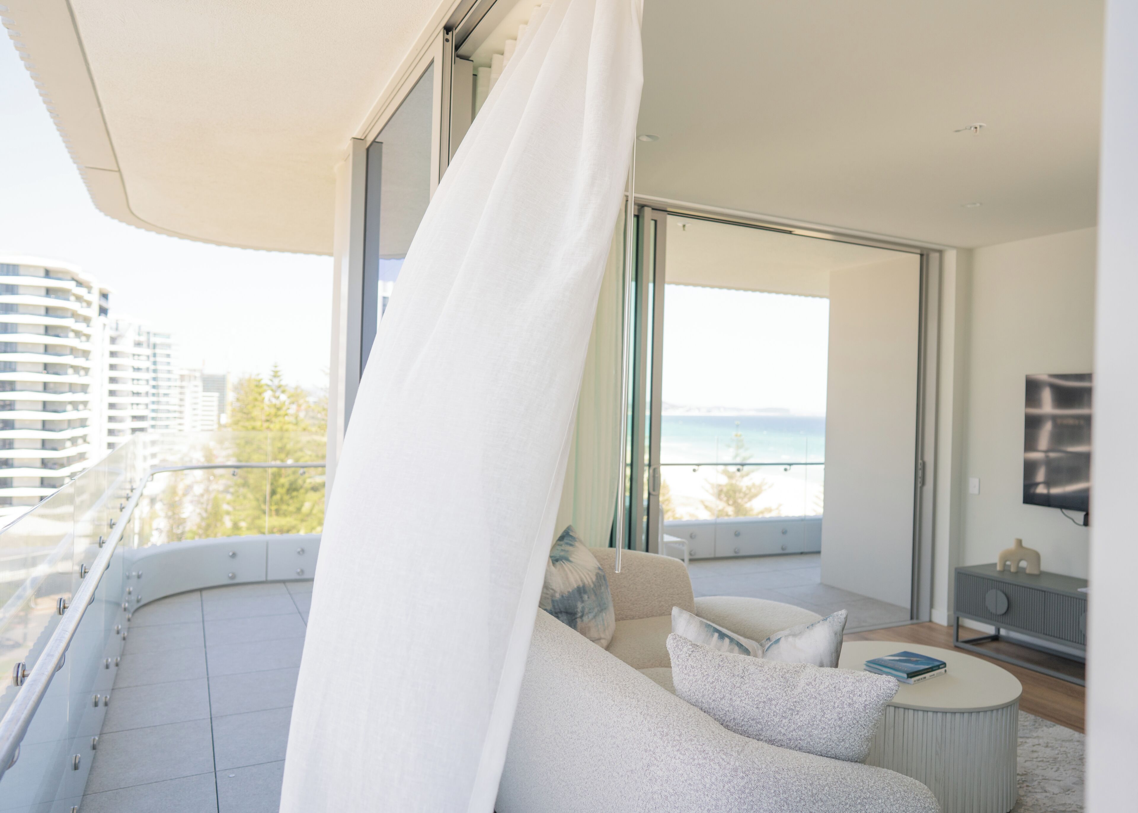 Un balcón moderno con barandilla de cristal curvo, un sofá blanco, una mesa de centro redonda y vistas al mar.