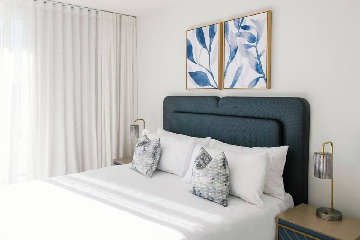 A neatly made bed with white bedding and patterned pillows, a dark headboard, and two wall-mounted artworks.