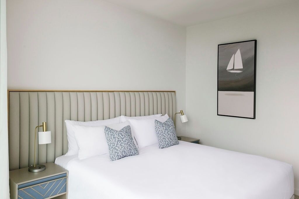A neatly made bed with white bedding and patterned pillows, a bedside table with a lamp, and a framed picture of a sailboat on the wall.