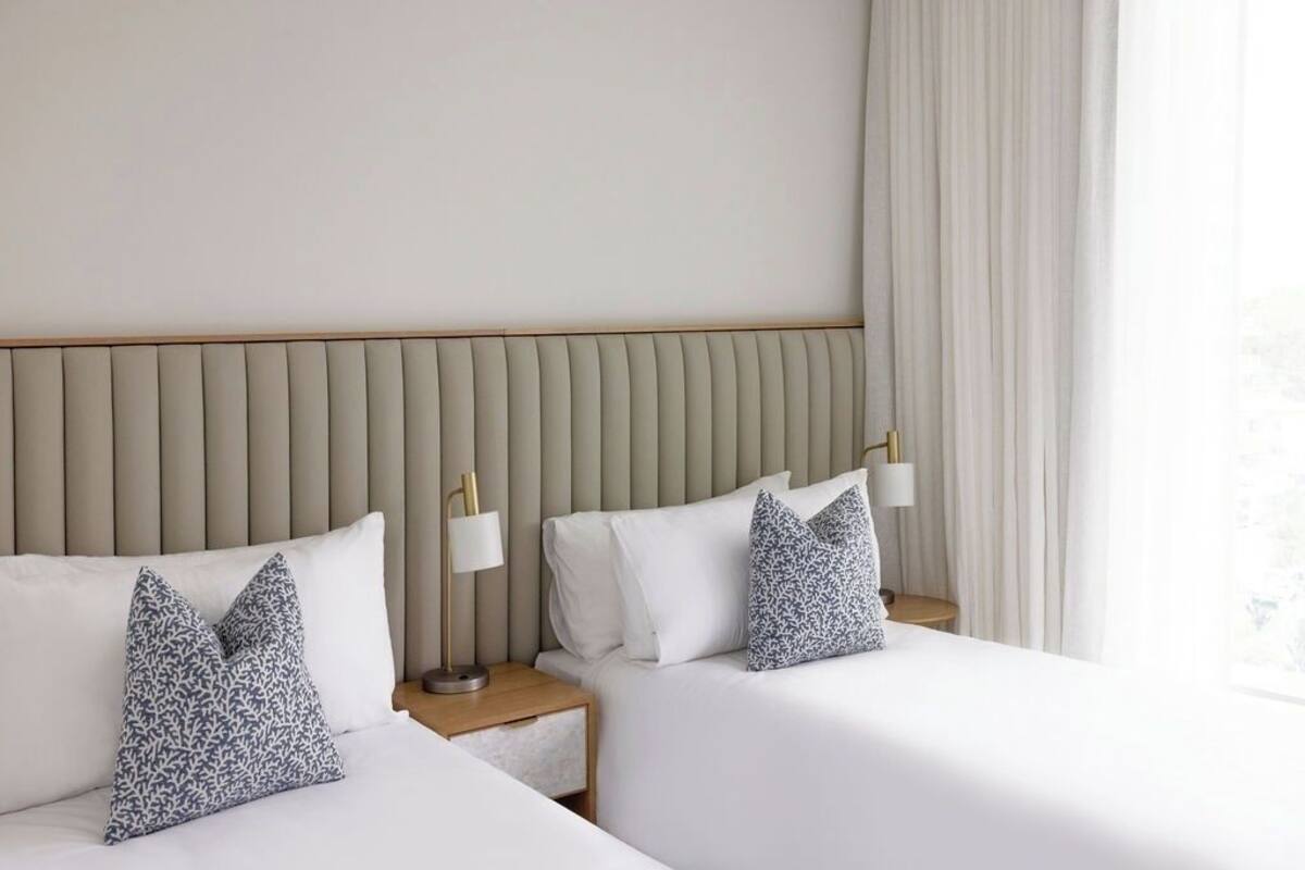 A hotel room with two beds, a headboard, bedside lamps, and a window with curtains.