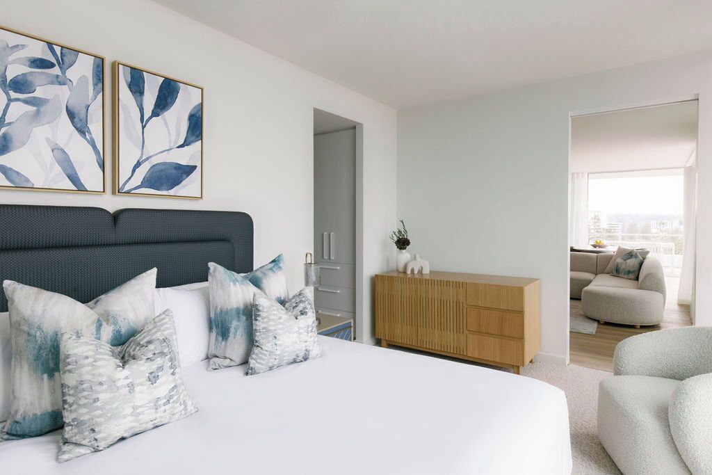 Une chambre à coucher moderne avec un lit, des oreillers, une commode en bois et une vue sur une autre pièce.