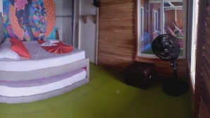 Een kamer met een bed, een kleurrijk schilderij, een ventilator en een houten vloer.