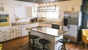 A modern kitchen with a central island, bar stools, a refrigerator, and a pendant light fixture. - Vibrant Forest Retreat | Game Room, Spa, Fire Pit - Sleeps10 (Spirit Lake)