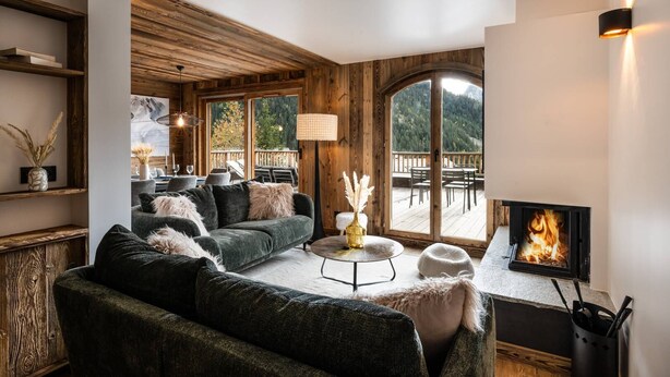 A cozy living room with a fireplace, wooden walls, and a large window overlooking a deck. - N°2: Beautiful 10 people apartment with sauna  et  south facing terraces (CHAMPAGNY EN VANOISE)