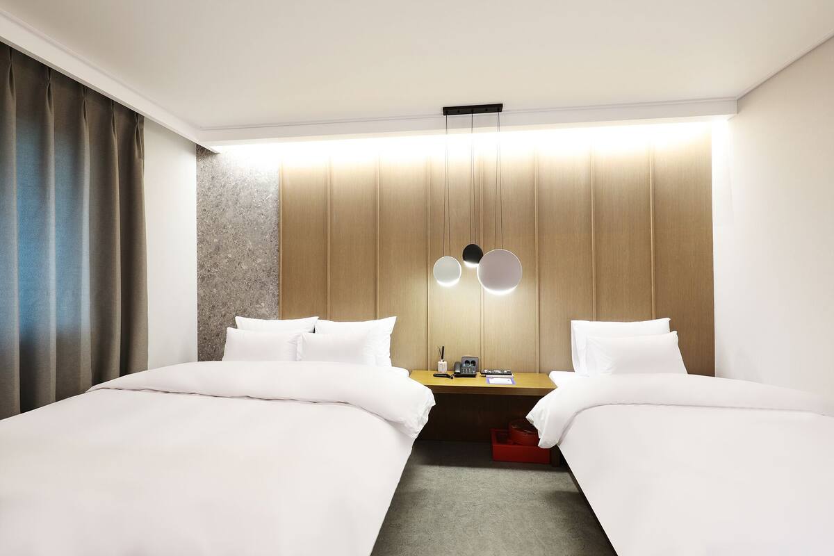 Une chambre d’hôtel avec deux lits, une tête de lit en bois, une table de chevet avec une lampe et une valise rouge posée sur le sol.