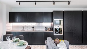A modern kitchen with dark cabinetry, a built-in microwave, and a dining area with a grey sofa and a table set for four. - Icon Tower Flat II (London)