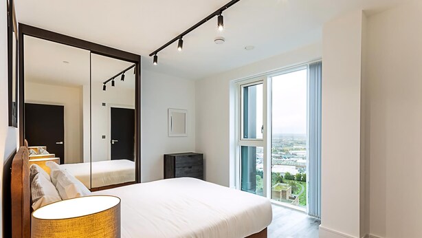 Ein modernes Schlafzimmer mit einem großen Bett, einem Spiegel, einem Nachttisch und Blick auf die Stadt durch ein großes Fenster.