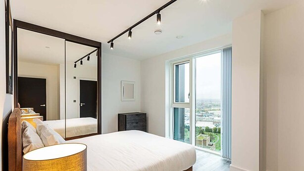 A modern bedroom with a large bed, a mirror, a bedside lamp, and a view of the city through a large window. - Icon Tower Flat II (London)