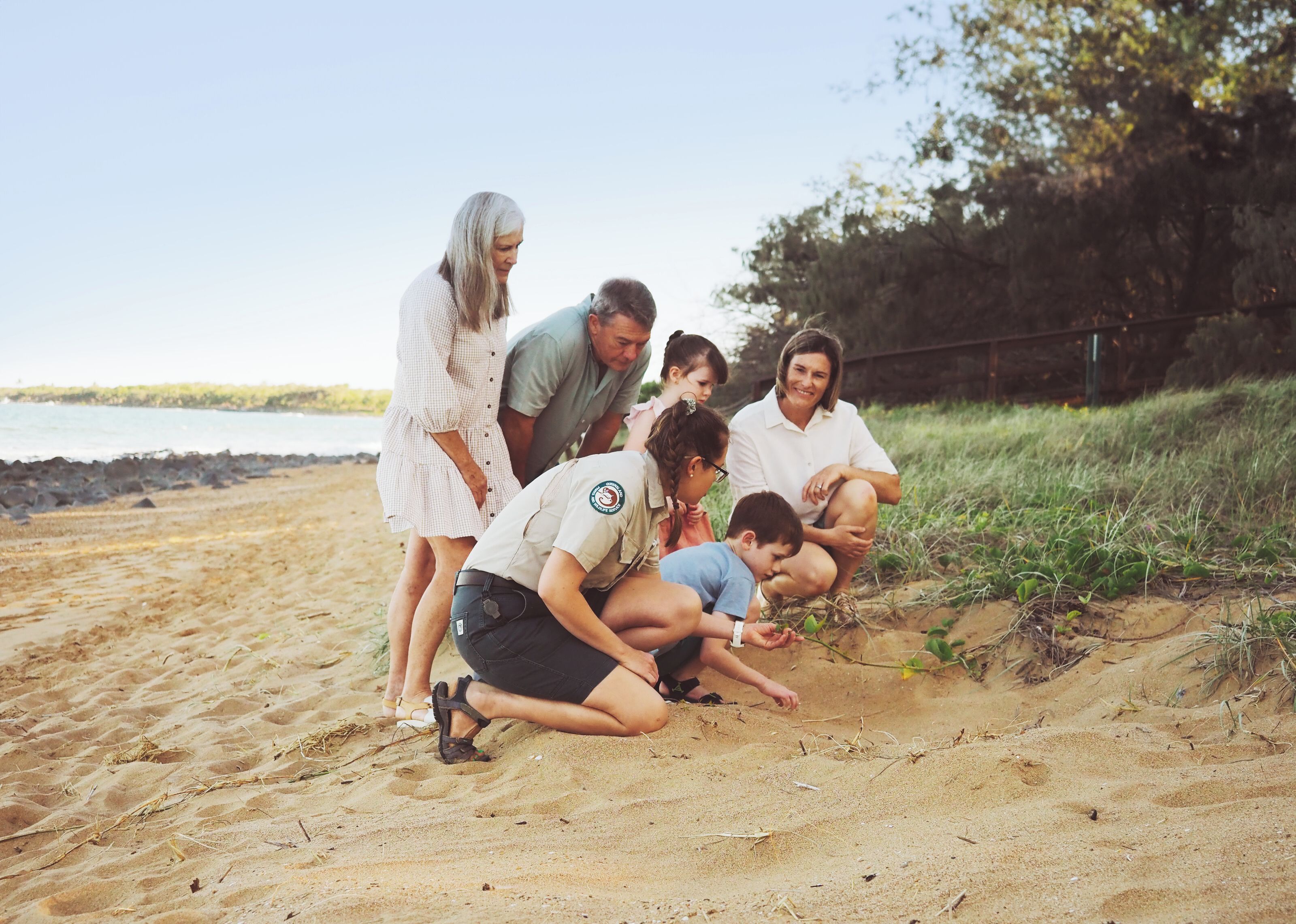 Een groep mensen op een zanderig strand, met een grasveld en bomen op de achtergrond.