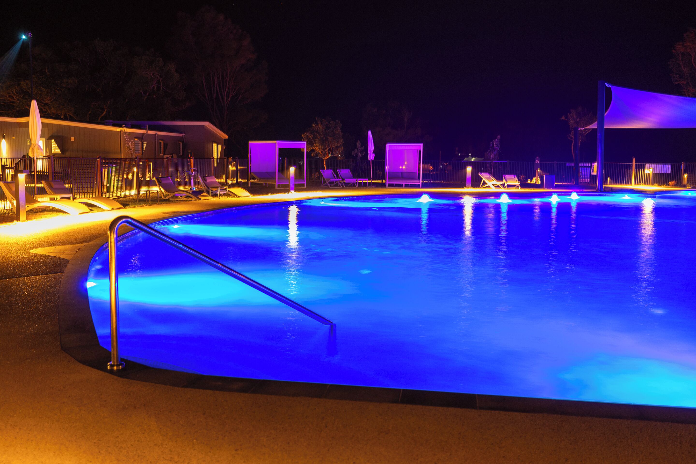 A nighttime swimming pool with blue lighting, lounge chairs, and a covered area.