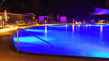 A nighttime swimming pool with blue lighting, lounge chairs, and a covered area.