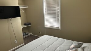 Ein Schlafzimmer mit einem Bett, einem an der Wand montierten Fernseher, einem Fenster mit Jalousien und einem Regal mit gefalteter Kleidung.