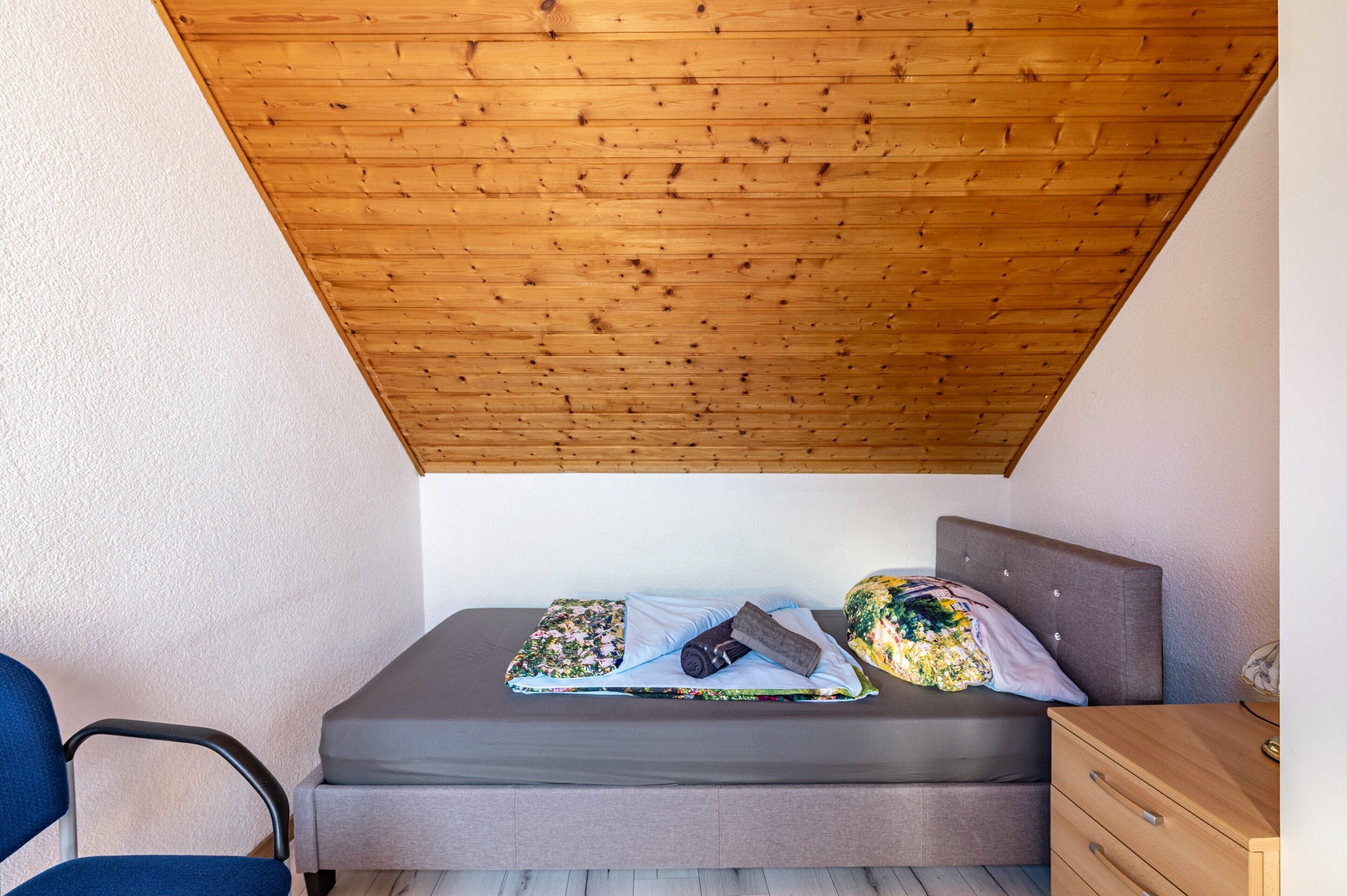A small bedroom with a sloped ceiling, a bed with a grey mattress, a wooden nightstand, and a chair.