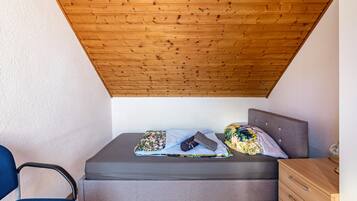 A small bedroom with a sloped ceiling, a bed with a grey mattress, a wooden nightstand, and a chair.