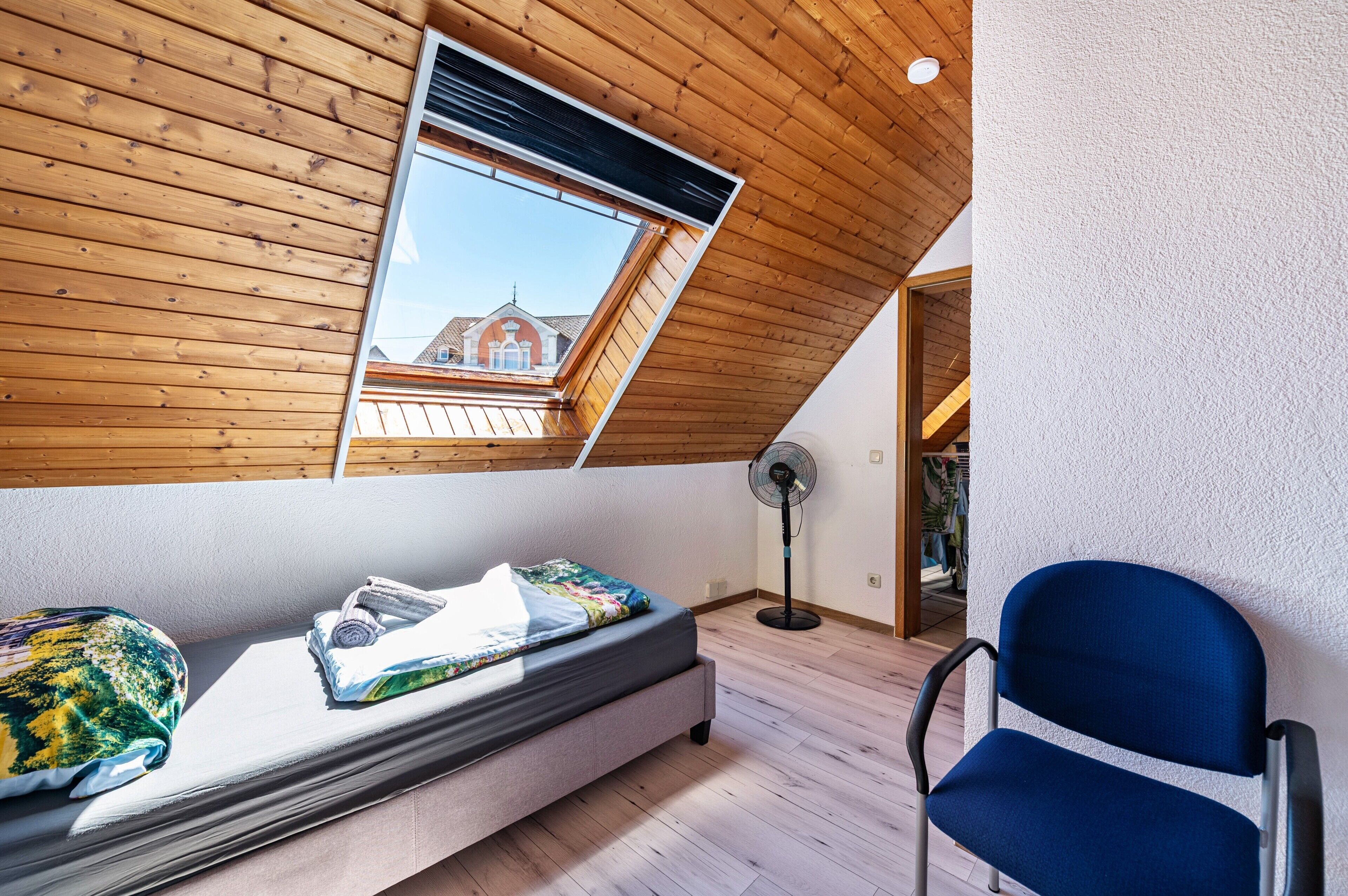Une chambre à coucher aménagée dans un grenier, avec un plafond en pente, un puits de lumière, un lit recouvert d’une couverture colorée, une chaise bleue et un ventilateur sur pied.