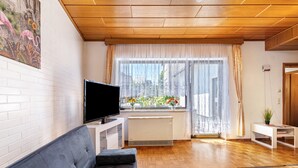 A living room with a sofa, a TV, a radiator, and a window with curtains. - Holiday Home 'Eiffelperle' with Private Terrace, Garden, Infrared Sauna & Wi-Fi (Kaisersesch)