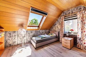 Un dormitorio con techo inclinado, tragaluz, cama, mesita de noche, ventilador y ventana con cortinas.