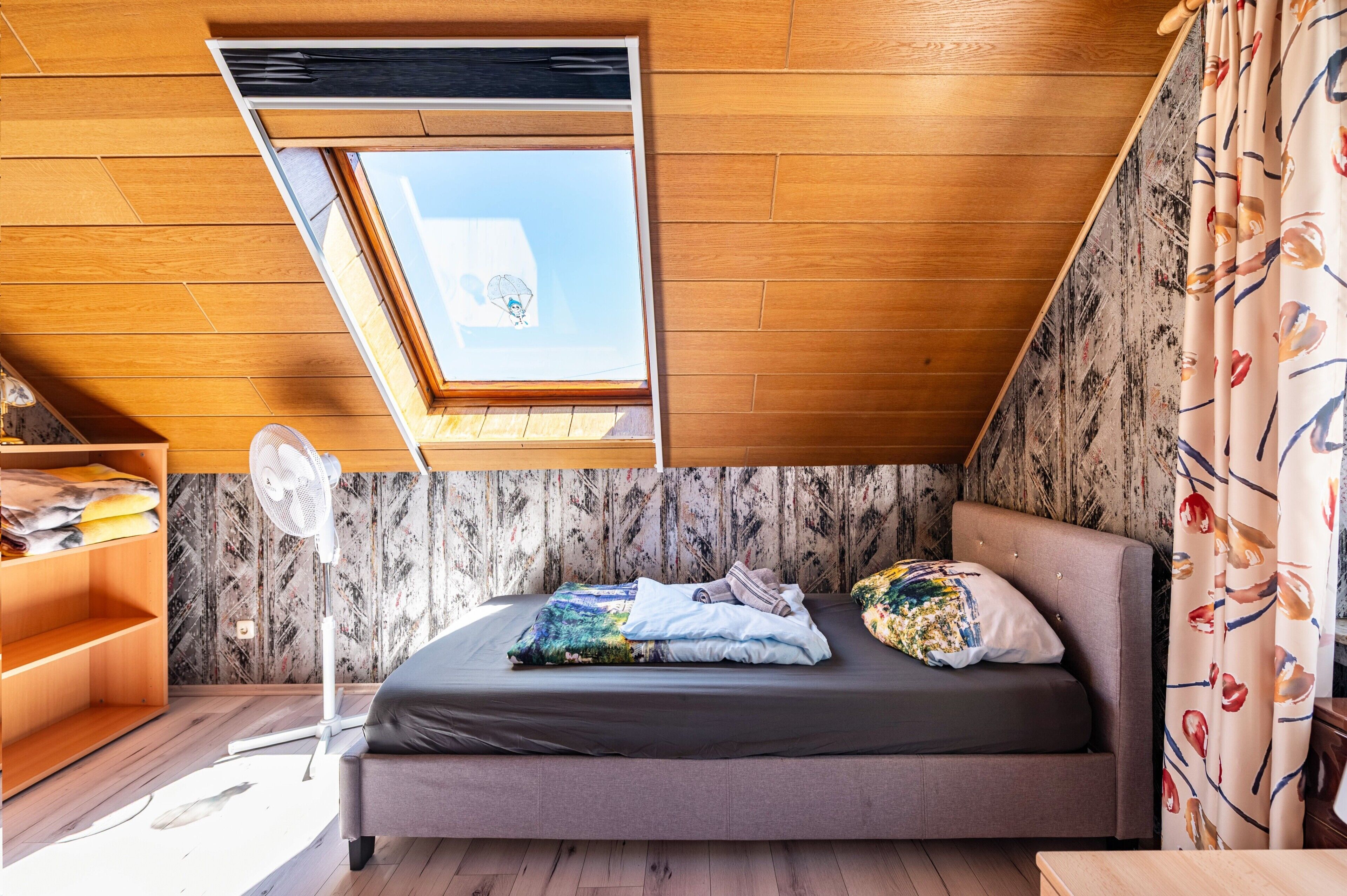 Une chambre avec un plafond en pente, un puits de lumière, un lit avec des oreillers, un ventilateur et une étagère en bois.