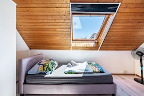 Quarto em estilo loft com teto inclinado, claraboia, cama com almofadas e um ventilador.