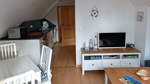 A modern kitchen with a microwave, toaster, and coffee maker. There is a dining area with a white table and chairs. A television is on a wooden stand.