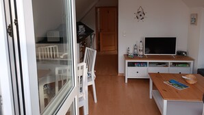 A room with a sliding glass door, a white chair, a wooden table, a TV, and a white cabinet with a shelf.