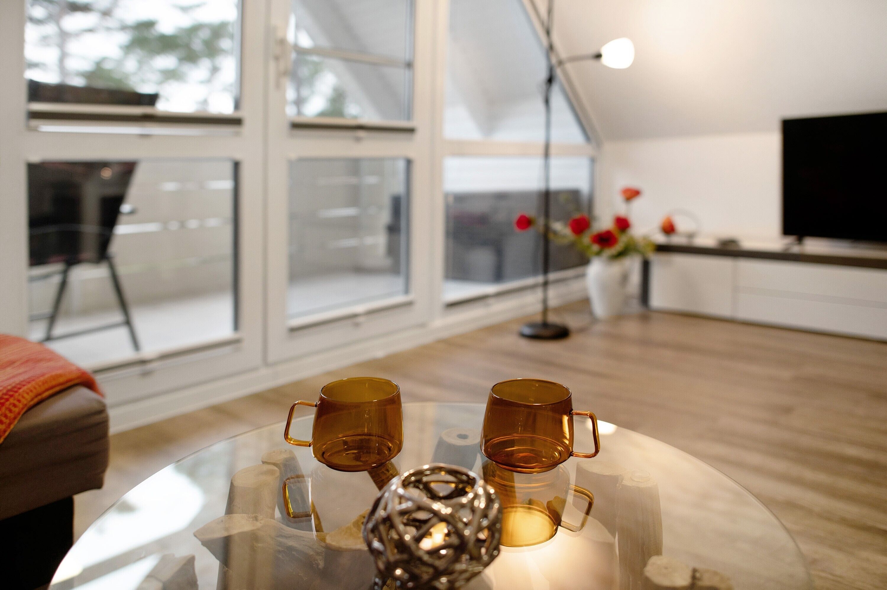 A modern living room with a glass coffee table, two brown cups, a decorative metal sphere, a vase with red flowers, and a large window.