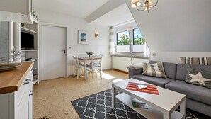 A modern living room with a sofa, a dining table with chairs, a kitchen area, and a large window with blinds. - Apartment 'Jule' with Shared Terrace, Shared Garden and Wi-Fi (Pellworm)