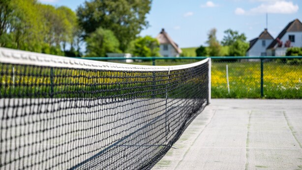 Een tennisbaan met een net, een groene hek en huizen op de achtergrond.