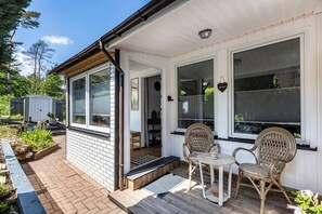 Eine überdachte Veranda mit zwei Korbstühlen und einem kleinen Tisch, eine Tür, die zu einem Innenbereich führt, und ein kleiner Gartenbereich mit Pflanzen.