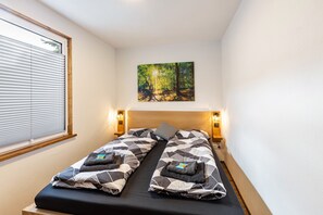 Ein Doppelbett mit geometrischem Bettbezug, zwei Kissen, Nachttischlampen, ein Gemälde von einem Wald und ein Fenster mit Jalousien.