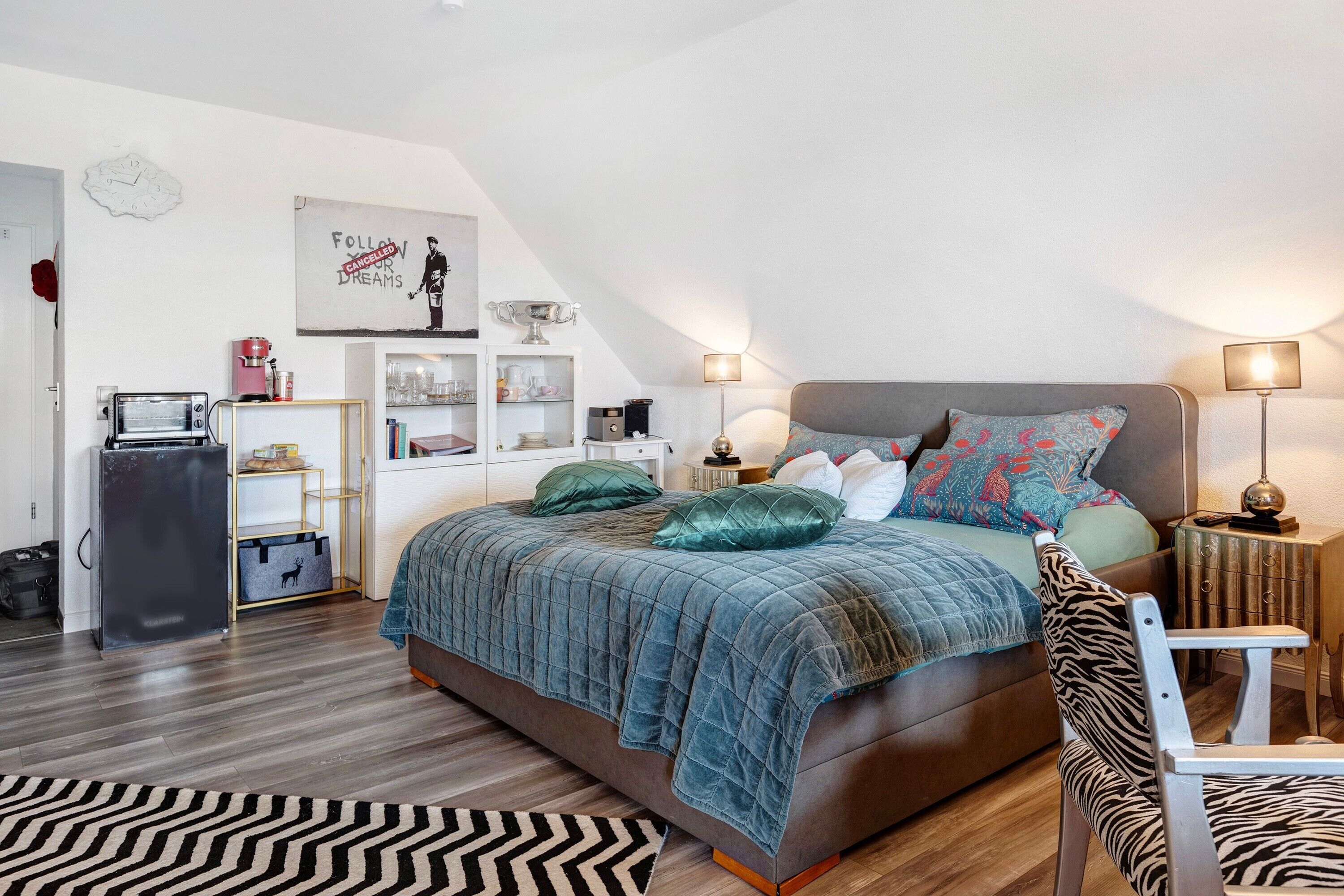 Ein Schlafzimmer mit einem Bett, einem Sessel, einem Schreibtisch, einer Lampe, einer Uhr und einem Regal mit verschiedenen Gegenständen.