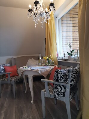 A dining area with a table, chairs, a chandelier, a window with blinds, and a plant on a windowsill.