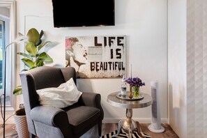A living room with a grey armchair, a round side table with a vase and a bottle, a flat-screen TV mounted on the wall, and a large poster with the text 'LIFE IS BEAUTIFUL'.