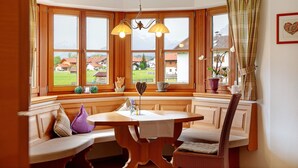 A corner dining area with a wooden table and bench, large windows with a view of houses and mountains, and a heart-shaped decoration on the windowsill. - Apartment 'Landhaus Bergkulisse' with Mountain View, Shared Terrace and Wi-Fi (Wallgau)